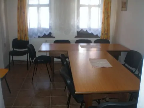 Jídelna s dlouhými stoly a židlemi, dvě okna se záclonami
